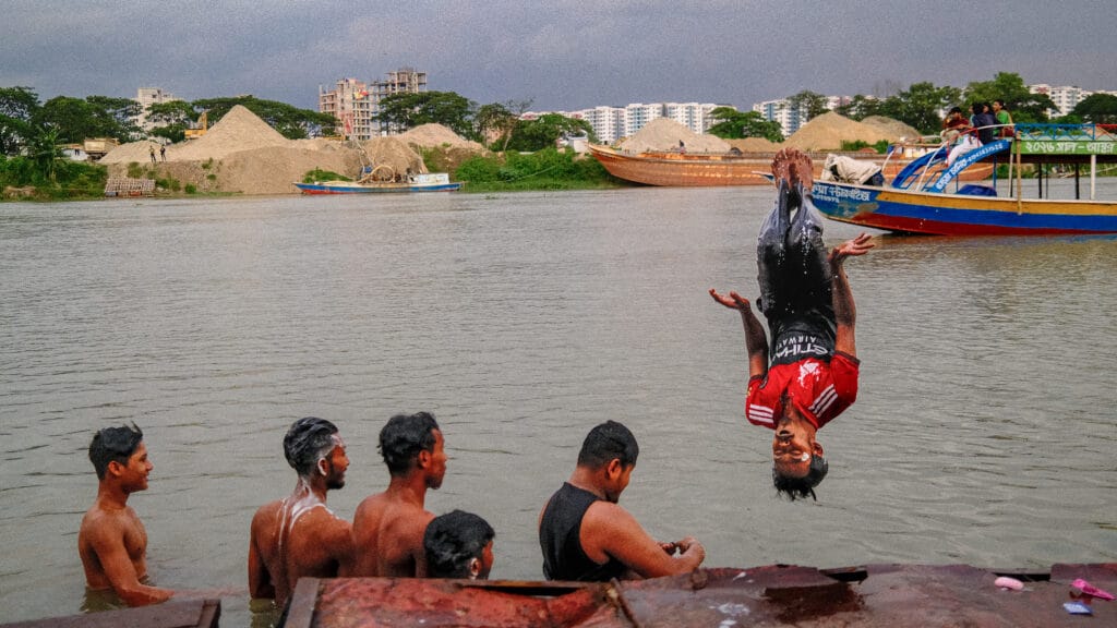 A young man flips above a river as friends watch, with boats, sand piles, construction sites, and Dhaka’s apartment blocks behind them.
Caption: